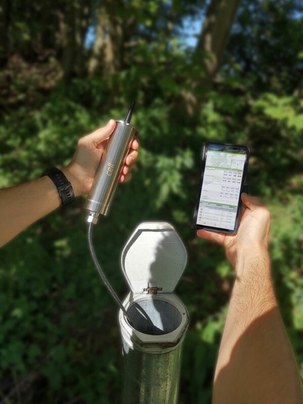 Person hält cell Datenlogger über geöffneter Grundwassermessstelle und liest aktuelle Messdaten am Smartphone aus.