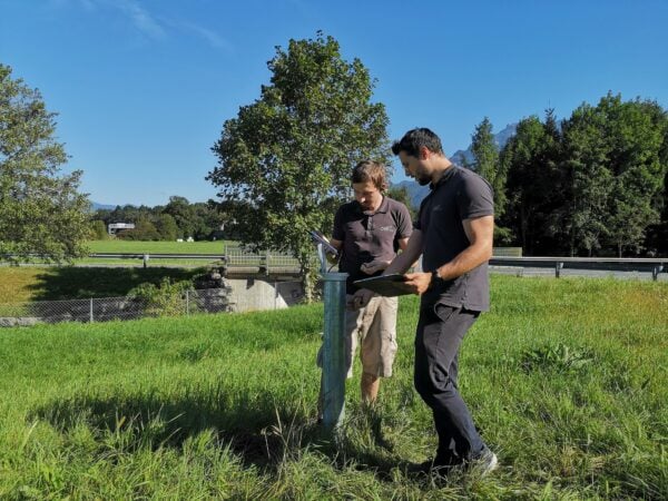 Zwei Techniker von cell analysieren eine Grundwassermessstelle im Gelände mit Tablet und Notizen bei sonnigem Wetter.